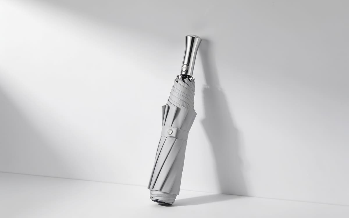 A neatly folded light grey umbrella with a silver handle on a light grey studio background