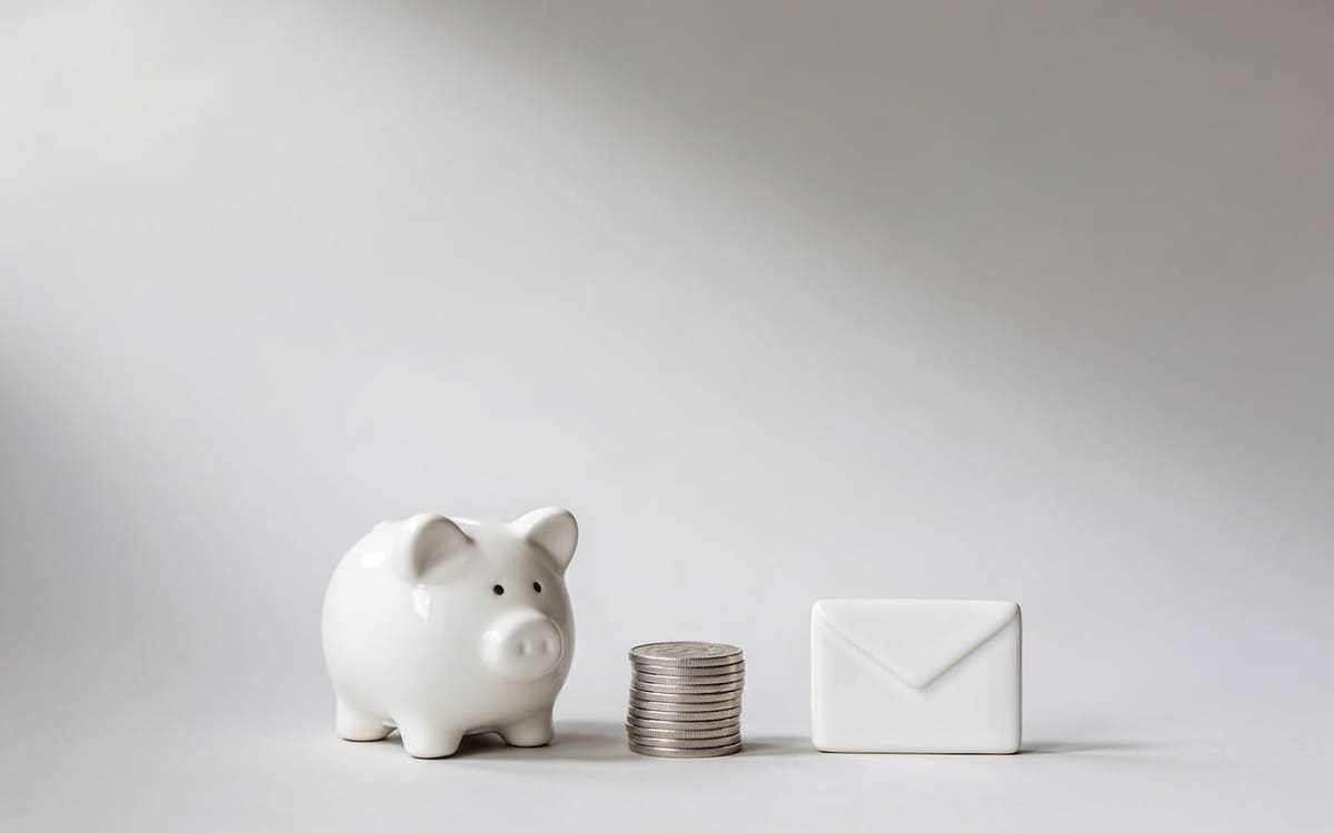 A white ceramic piggy bank beside silver coins and a blank envelope, on a light grey studio background