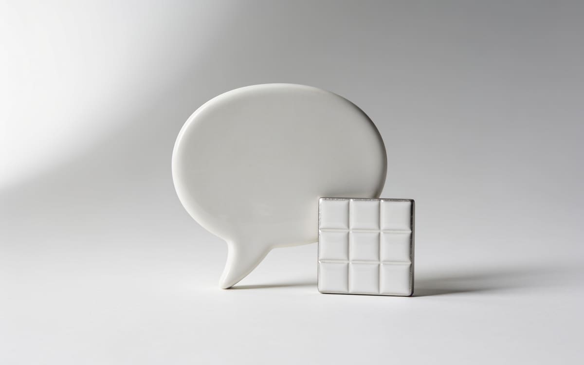 A white ceramic speech bubble beside a small tile with a square grid relief, on a light grey studio background